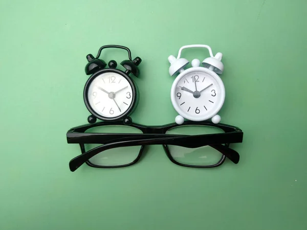 Black and white alarm clock and glasses on a green background.