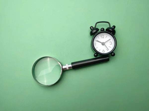 Black alarm clock and magnifying glass on a green backgrounf.