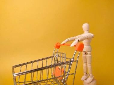 Ahşap manken sarı arka planda alışveriş arabası tutuyor..