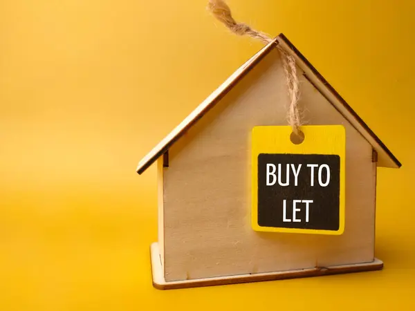 Wooden house and wooden board with text BUY TO LET on a yellow background.