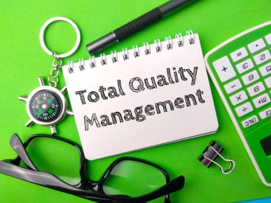 Üstteki görünüm pusulası, hesap makinesi, kalem ve not defteri ile yeşil arkaplan üzerinde toplam Kalite Yönetimi.