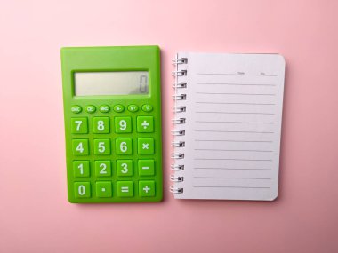 Üst görünüm yeşil hesap makinesi ve boş not defteri pembe arkaplanlı bir ofis masasının üzerinde. İş konsepti.