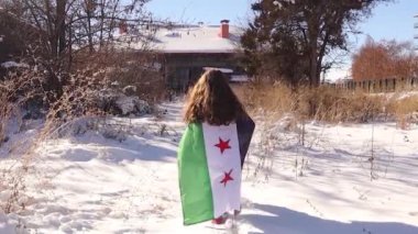 Küçük Suriyeli kız, umut ve dayanıklılığı simgeleyen devrim bayrağına sarılı olarak karda yürüyor. Yolculuğu kendisi ve tüm Suriyeliler için daha parlak bir gelecek hayallerini yansıtıyor..