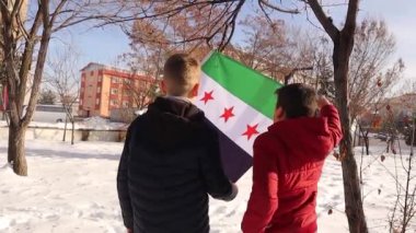 İki çocuk, kendileri ve sürgündeki tüm Suriyeliler için daha iyi bir gelecek umuduyla, karlı bir atmosferde vatanlarına dönmeyi hayal ederek, Suriye Devrim Bayrağına sarılıp bakıyorlar.