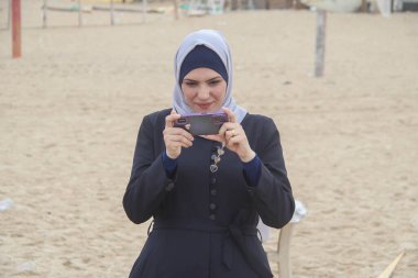 October 02, 2023 - Gaza, Palestine: Palestinian woman using phone while enjoying the winter weather on the Gaza City sea daily life.   clipart