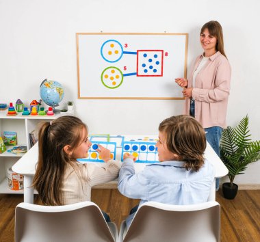 Bir öğretmen ve iki çocuk etkileşimli eğitim araçları kullanarak bir sınıfta matematik öğreniyorlar. Çocuklar elle sayım faaliyetiyle meşgulken, öğretmen konseptleri açıklarken...