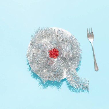 Gümüş parlak çelenk, beyaz yuvarlak tabakta kırmızı kurdele ve parlak mavi arka planda çatal içeren minimal Noel masası düzeni. Yaratıcı yeni yıl ya da Noel konsepti. Şenlikli yemek retro gerçeküstü fikir