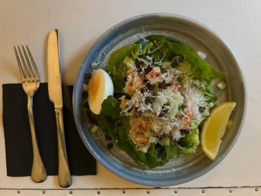 Kerevit ve marul yapraklı karides deniz ürünleri salatası, haşlanmış yumurta, tere, kapari, salatalık ve limon dilimleri bıçak çatallı düz bir peçete masasında servis edilir.