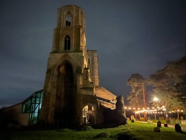 Night time landscape of Wymondham abbey in Norfolk East Anglia lit up Winter evening showing the ruined West Tower and ancient stone building with refurbished new glass build in the graveyard setting