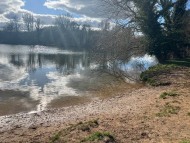 Landscape of Norfolk Broad with sandy shore by large lake , the Spring blue sky with sun light through white puffy cloud and trees reflected  in calm river water on bright cold day in East Anglia UK