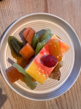 Lokantadaki gri tabakta renkli meyveli tatlının üzerine bir dilim tatlı peynirli puding ve olgun sulu kivi, kavun, böğürtlen ve mago renkli parlak renkli meyveler.