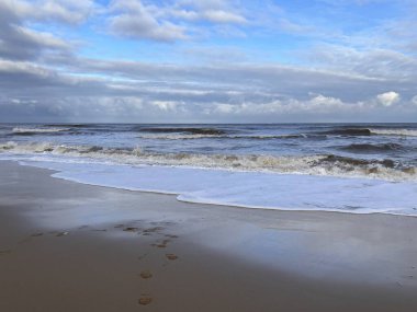 Göz kamaştırıcı, vahşi kumlu plaj manzarası beyaz dalgalarla okyanus suyundaki bulutları yansıtıyor engin deniz kıyısına karşı ve gri gri bulutlu gri gökyüzü kış günü