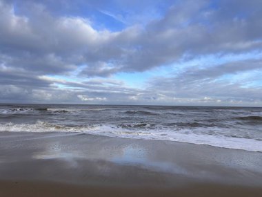 Göz kamaştırıcı, vahşi kumlu plaj manzarası beyaz dalgalarla okyanus suyundaki bulutları yansıtıyor engin deniz kıyısına karşı ve gri gri bulutlu gri gökyüzü kış günü