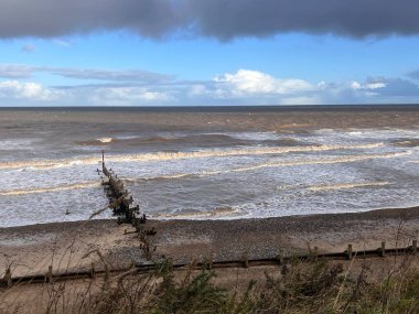 Landscape of beautiful sandy beach on stormy cloudy day with wood pier groyne structure looking to ocean horizon with large surf waves and bleu grey dramatic clouds in Winter in East Anglia  clipart