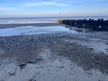 Beautiful beach landscape in Winter at sun down with blue clear sky and wood groyne sea defence looking out to ocean with skies and cloud  reflected in salt water pool on sany shore with pebbles clipart