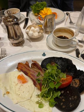 İngiliz kahvaltısı, kızarmış yumurta, pastırma, siyah puding, sosis, domates mantarı, fincan kahve. Ve gümüş kaplar ve tereyağlı çatal bıçak seti otel restoranında beyaz keten masa örtüsünün üzerinde.