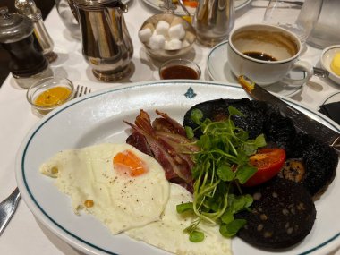 İngiliz kahvaltısı, kızarmış yumurta, pastırma, siyah puding, sosis, domates mantarı, fincan kahve. Ve gümüş kaplar ve tereyağlı çatal bıçak seti otel restoranında beyaz keten masa örtüsünün üzerinde.