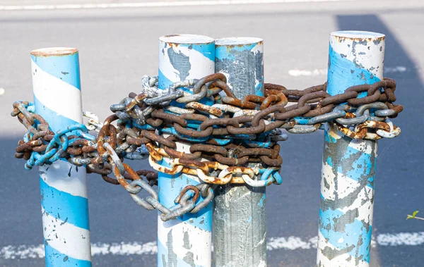Old rusted industrial steel chain wrapped around several metal parking poles painted blue and white