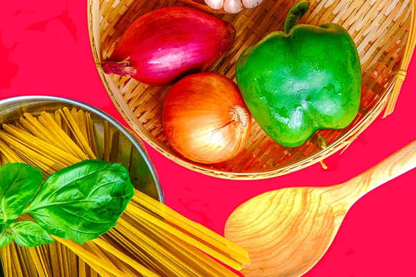 Flat lay of green pepper and onions in bamboo basket and uncooked pasta in sauce pan next to wood spoon on red background.