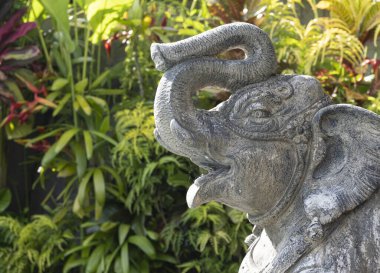 Gövdesi bükülmüş, başının üstünde taş bir fil. Arka planda tropik yeşil bitkiler var. Bali, Endonezya. Boşluğu kopyala