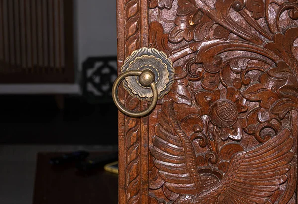 Balinese carved wood door with brass door knob ring and partially open darkened interior space. Space of text