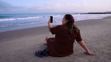 Önlüklü yaşlı bir kadın denizin önünde sandaletlerinin yanında oturmuş Kolombiya, Cartagena de Indias sahillerinde selfie çekiyor..