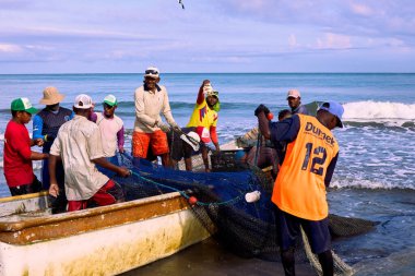 Cartagena, Bolivar Kolombiya - 1 Şubat 2024: Kanoyu çevreleyen ve ağları toplamayı ve deniz kıyısındaki balıkları ayırmayı bitiren bir grup balıkçı. Balıkçılardan biri memnuniyet işareti olarak bir balık gösteriyor.