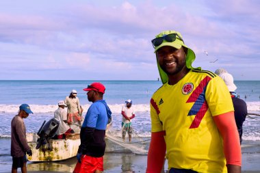 Cartagena, Bolivar Kolombiya - 1 Şubat 2024: Ön planda, siyah bir balıkçı kameraya ve arka planda, motorlu bir tekne ve arkadaşları günlük balıkçılık araçlarını tamir ettikten sonra,