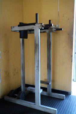 Halter tutacağı olan metal bir halter rafı ve yerde ağırlıklar, spor salonunda sarı bir duvara dayanıyor..