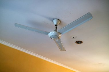 A ceiling fan with three blades mounted on a white ceiling, with a yellow wall in the background. The fan appears to be slightly dusty, indicating it may not have been used recently.