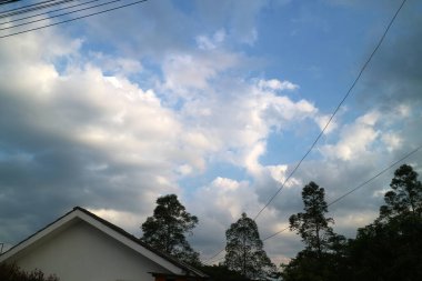 Parçalı bulutlu bir gökyüzü manzarası mavi yamalar ve dağınık bulutlar, ağaçların siluetleriyle çerçevelenmiş ve ön planda bir ev.