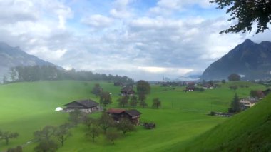 İsviçre Alplerinde Yeşil Tarlalar ve Dağ Manzaraları. İsviçre Alpleri 'ndeki uzak dağlarla yeşil tarlalardan oluşan sakin bir manzara. Çiftlik evleri kırsal kesimde, sonbahar renkleri bulutlu gökyüzünün altında belirmeye başlıyor..