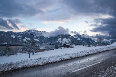 Yoğun kar yığınlarıyla çevrili yeni sürülmüş bir yolun yer aldığı pitoresk bir kış manzarası. Uzakta, sakin dağlar ve sakin bir göl dramatik bulutlar ve karla kaplı ağaçlarla çevrelenir..
