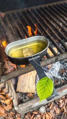 Kamp ateşi ızgarasının üzerinde kaynayan suda demlenen bir çay poşetiyle metal bir tava. Duman ve alevler bu dingin vahşi doğada pişirme anını vurgular..