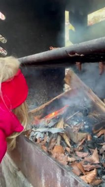 Soğuk hava için giyinmiş bir çocuk dikkatle kamp ateşine üflüyor ve büyümesine yardım ediyor. Kuru yapraklar ve sakin bir orman açık havanın doğal cazibesini ön plana çıkarıyor..