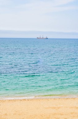 Uzakta bir kargo gemisi olan engin mavi bir deniz manzarası. Turkuaz su yumuşak bulutlu bir gökyüzünün altında kumlu sahille nazikçe buluşur ve huzurlu bir atmosfer yaratır..
