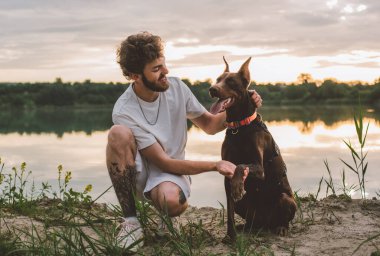 Genç adam güzel gün batımında gölün kenarında sevimli doberman köpeğiyle oynuyor.