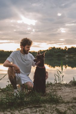 Genç adam güzel gün batımında gölün kenarında sevimli doberman köpeğiyle oynuyor.