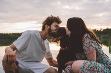 Genç mutlu çift piknik yapıyor ve gün batımında göl kenarında doberman köpekleriyle oynuyorlar.