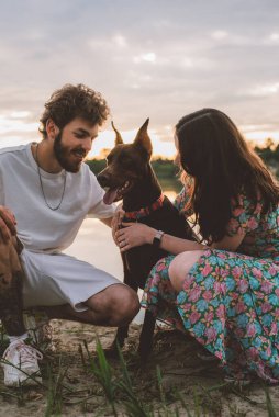 Genç mutlu çift piknik yapıyor ve gün batımında göl kenarında doberman köpekleriyle oynuyorlar.