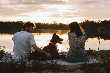 Genç mutlu çift piknik yapıyor ve gün batımında göl kenarında doberman köpekleriyle oynuyorlar.