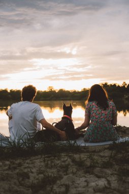 Genç mutlu çift piknik yapıyor ve gün batımında göl kenarında doberman köpekleriyle oynuyorlar.