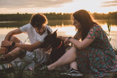 Genç mutlu çift piknik yapıyor ve gün batımında göl kenarında doberman köpekleriyle oynuyorlar.