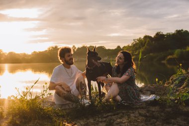 Genç mutlu çift piknik yapıyor ve gün batımında göl kenarında doberman köpekleriyle oynuyorlar.