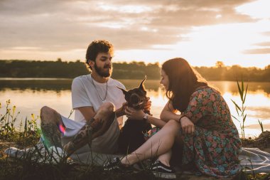 Genç mutlu çift piknik yapıyor ve gün batımında göl kenarında doberman köpekleriyle oynuyorlar.