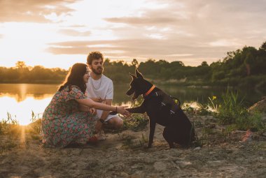 Genç mutlu çift piknik yapıyor ve gün batımında göl kenarında doberman köpekleriyle oynuyorlar.