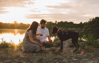 Genç mutlu çift piknik yapıyor ve gün batımında göl kenarında doberman köpekleriyle oynuyorlar.
