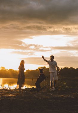 Genç mutlu çift piknik yapıyor ve gün batımında göl kenarında doberman köpekleriyle oynuyorlar.