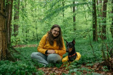 Genç bir kadın ormanda doberman köpeğiyle yürüyor. Açık havada oynayan sarı yağmurluk giyen kız ve köpek. İnsan ve köpek arkadaşlık kavramı