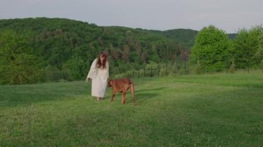 Genç ve güzel bir bayan köpeğiyle arka bahçede muhteşem bir tepe manzarasıyla oynuyor.